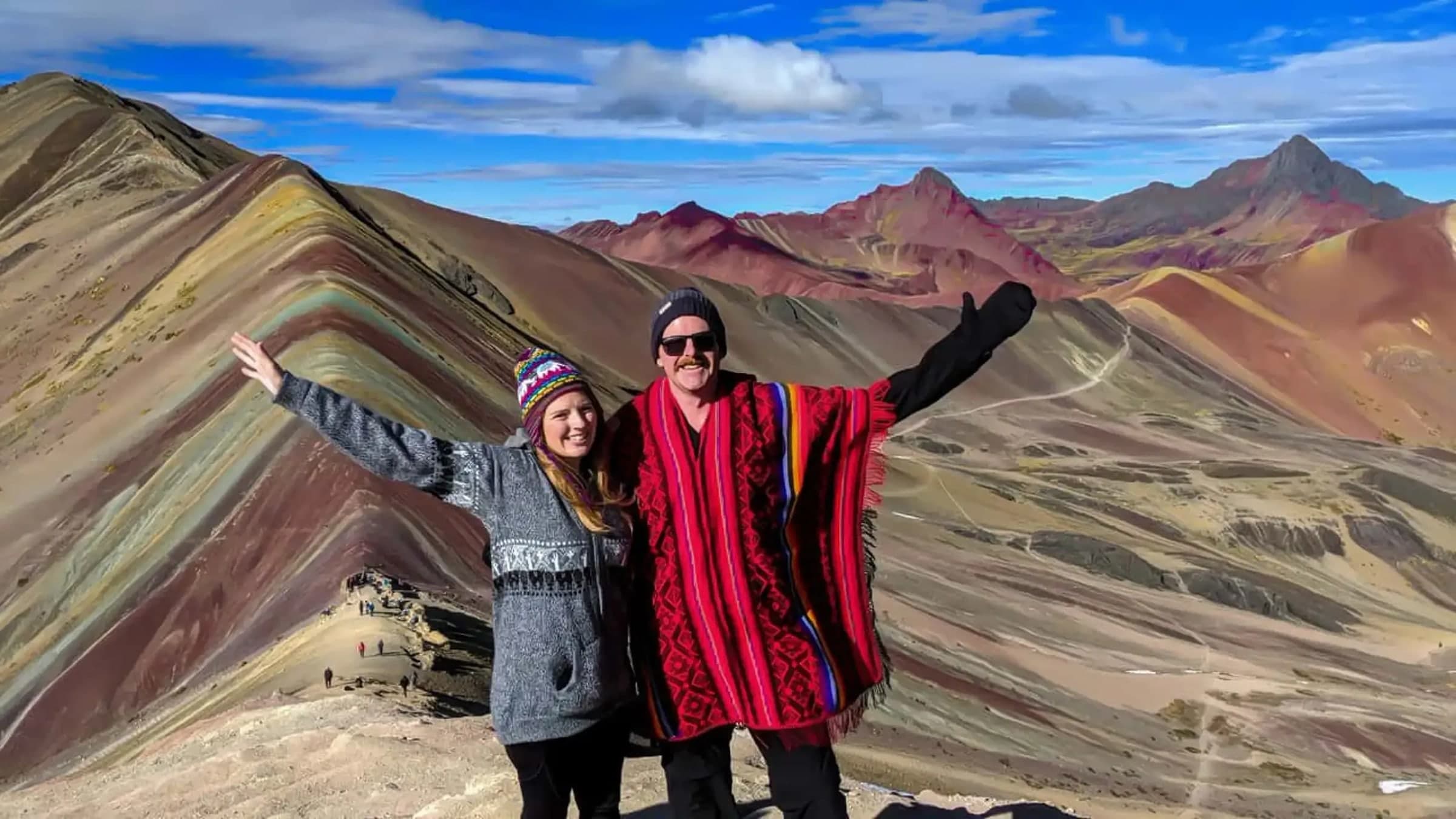 Two travellers in Andean ponchos in front of the coloured bands of Vinicunca